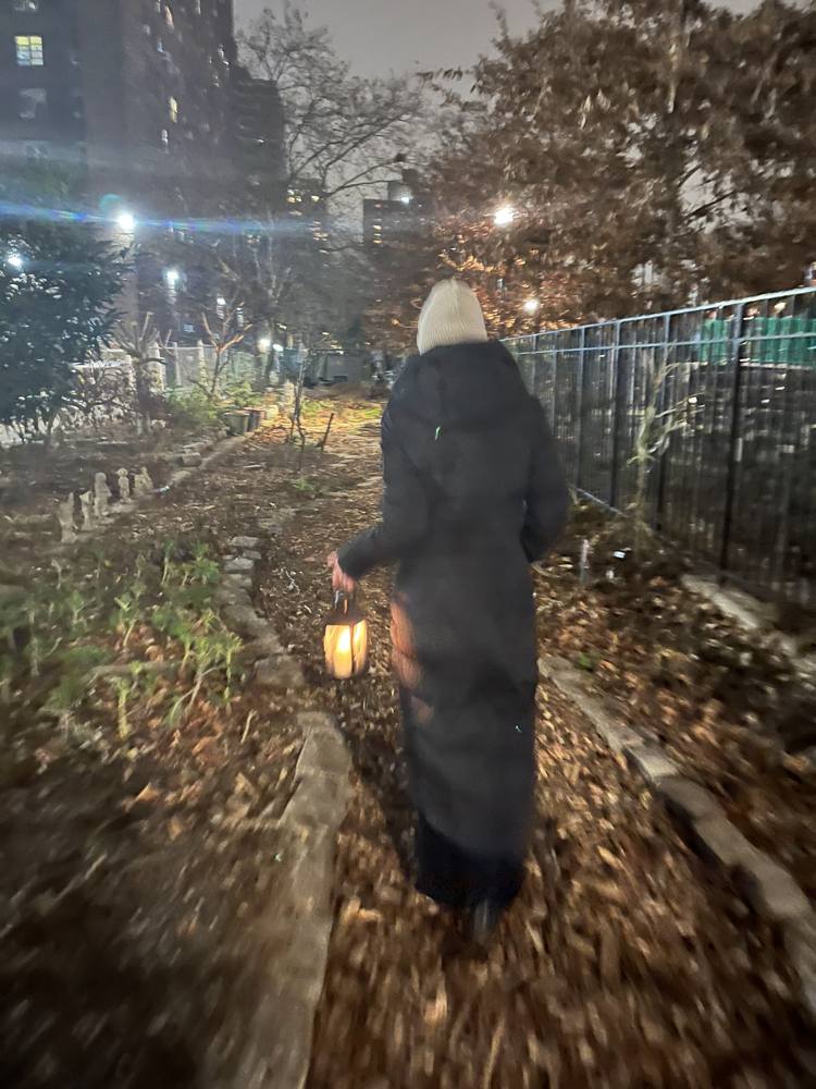 A figure in a long black coat carrying a lit lantern through a night garden