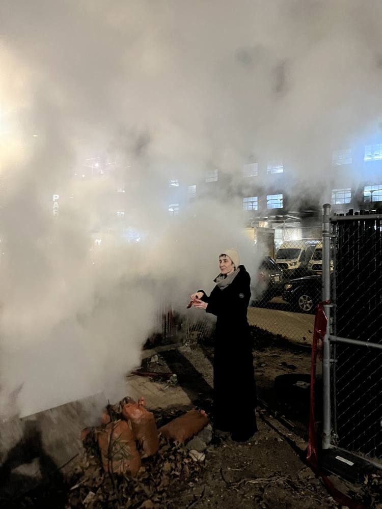 A figure in black making offerings beside a column of steam at night, Descent Ritual, New York City