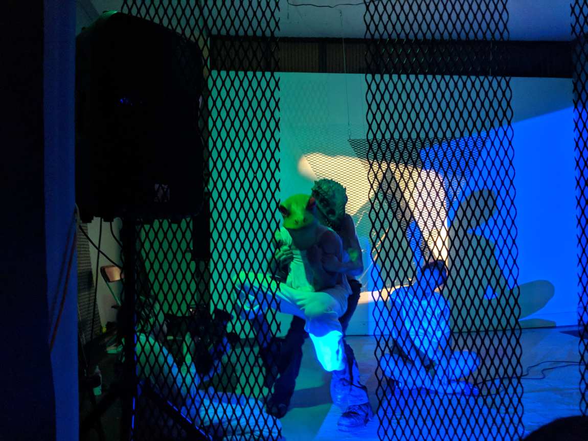 A masked reptilian figure carrying another performer, seen through the black lattice in green light