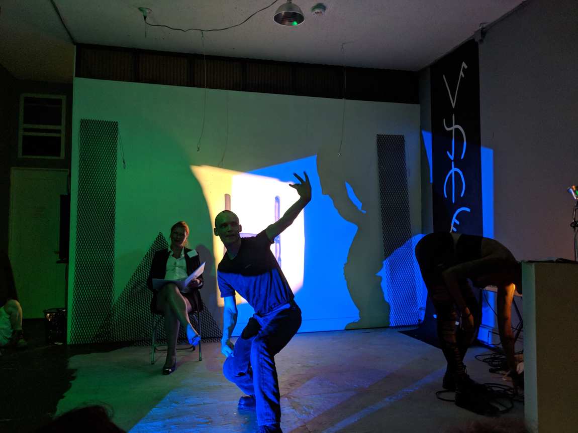 A performer crouching mid-gesture in blue light, casting a large shadow, a seated note-taker behind