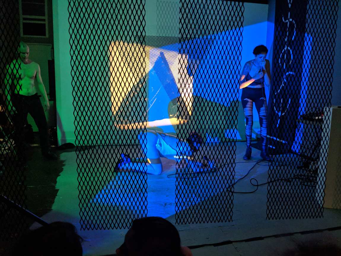 A shirtless figure at left and a performer at a microphone at right, behind hanging black lattice panels in blue and green light