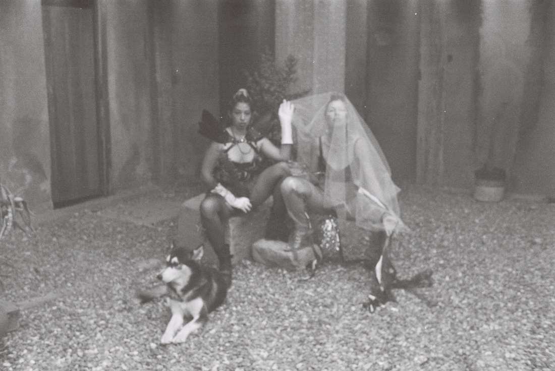 Black and white film photograph of two seated personas, one veiled, a husky lying on the gravel before them