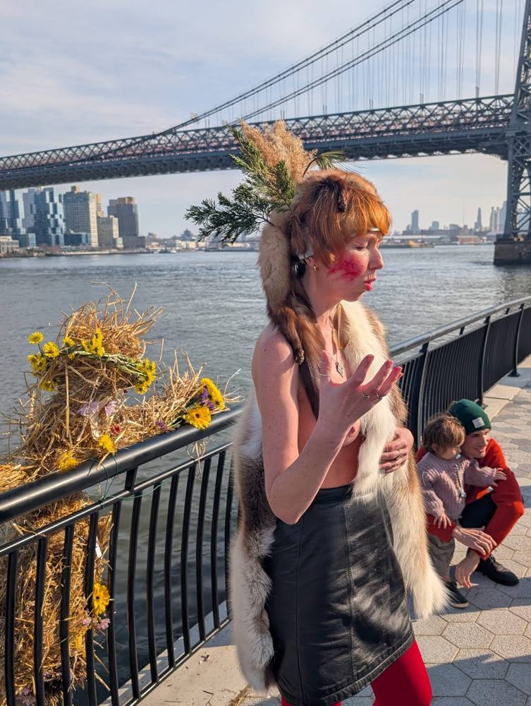 Dasha in a fur cape blessing the flower-dressed straw effigy at the East River railing, the Williamsburg Bridge behind