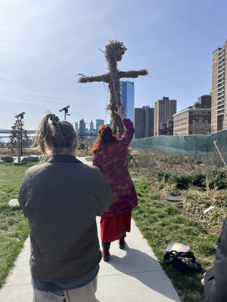 Dasha in a patterned red coat carrying the tall straw effigy overhead through the park, Manhattan skyline ahead