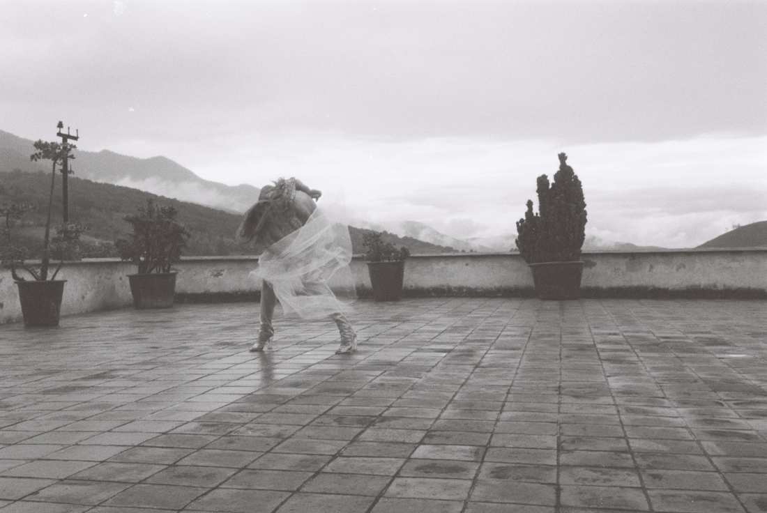 Film photograph of a figure bent deeply forward on the wet terrace, gathering the veil in one hand