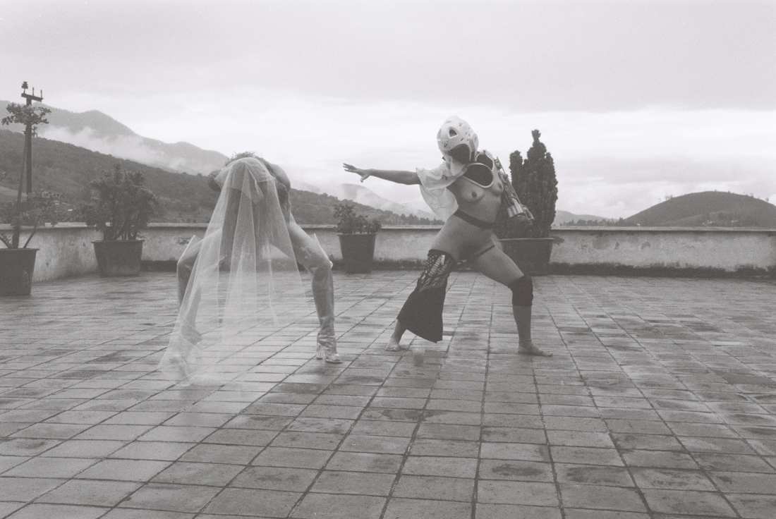 Film photograph of the helmeted figure reaching one arm toward the bent, veiled figure, mountains dissolving in cloud