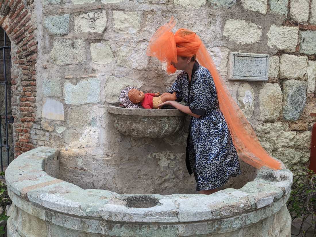 In an orange tulle veil and leopard robe, bathing a plastic doll in a stone wall fountain