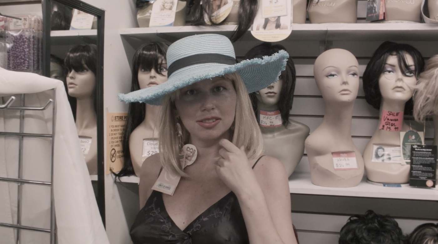 Natasha in a black slip and a fringed pale-blue sun hat, surrounded by wig-shop mannequin heads and price tags