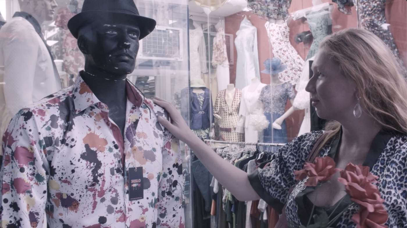 Natasha in a leopard-print top resting her hand on a black-faced mannequin in a fedora and paint-splattered shirt, in a crowded clothing shop window
