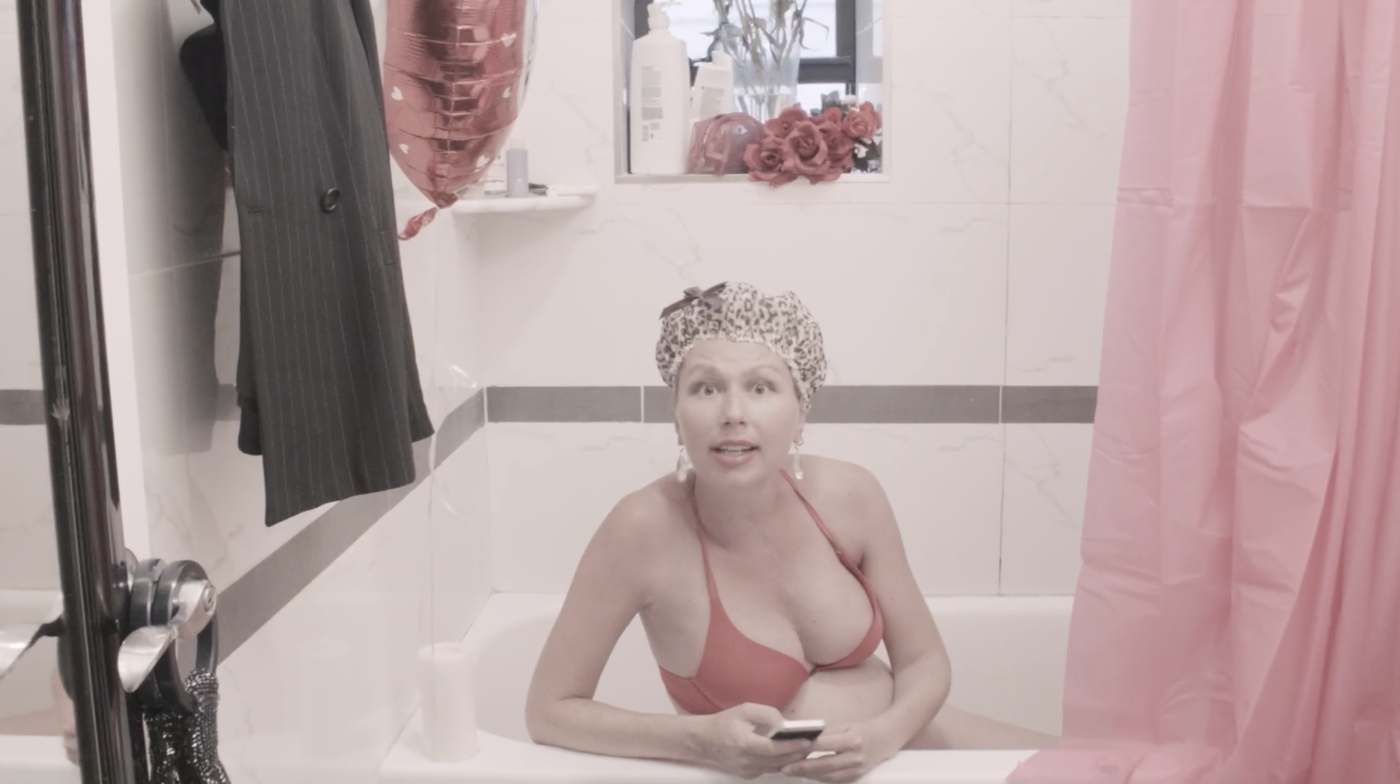 Natasha in the bathtub in a shower cap and coral bikini, phone in hand, addressing the camera, a red heart balloon and roses behind her