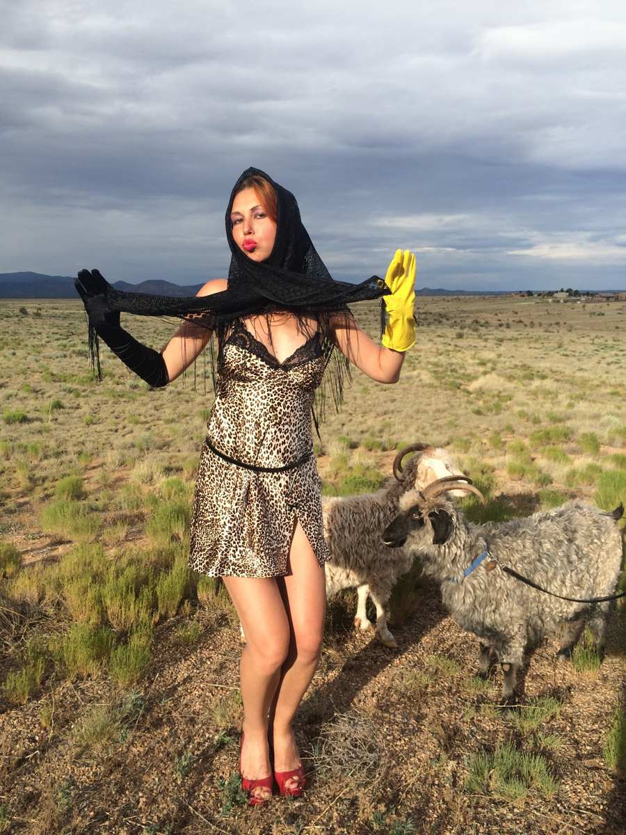 Natasha in the leopard slip with a black fringed shawl pulled over her head, blowing a kiss, goats grazing behind