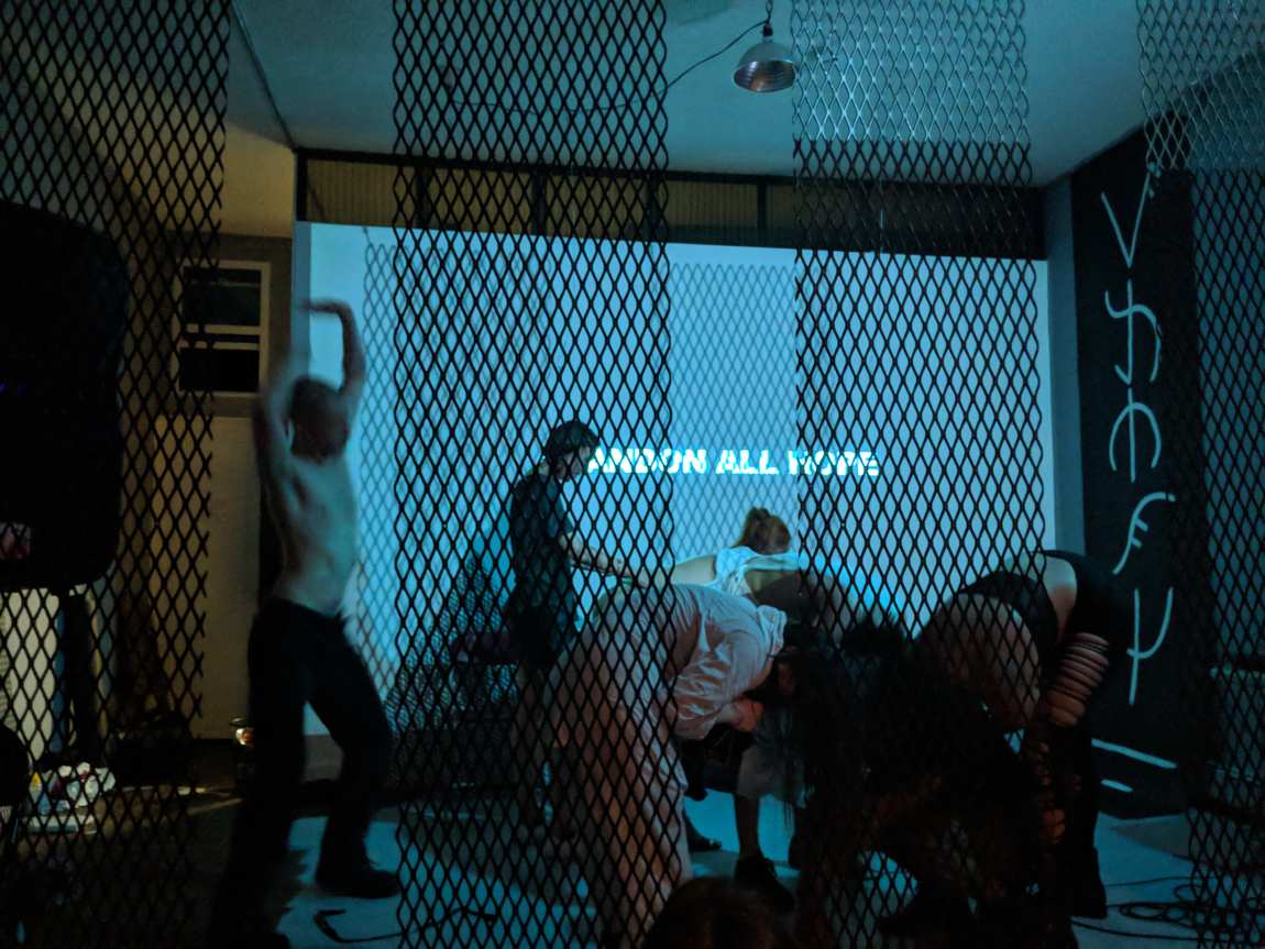Performers moving behind a chain-lattice screen, the projected words ABANDON ALL HOPE on the wall