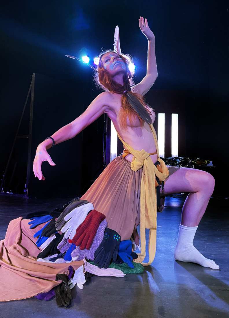 Performing in a skirt sewn from dozens of lost gloves, feathers in the hair, arm raised to the light