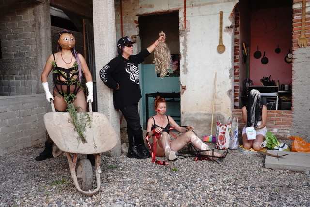 Personas assembling in a ruined brick courtyard: a masked figure with a wheelbarrow, a figure holding up a blond wig, performers preparing