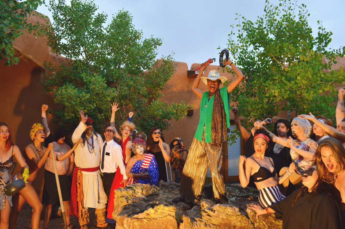 Personas gathered in an adobe courtyard at dusk, fists raised, one figure standing on a rock holding a pipe wrench and tambourine aloft