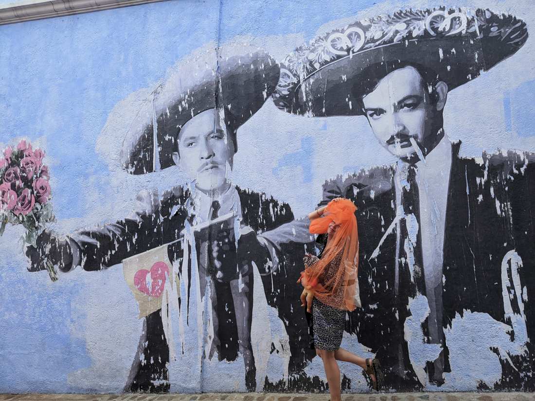 Posing playfully at the mariachi mural, orange veil pulled over the face, doll under one arm