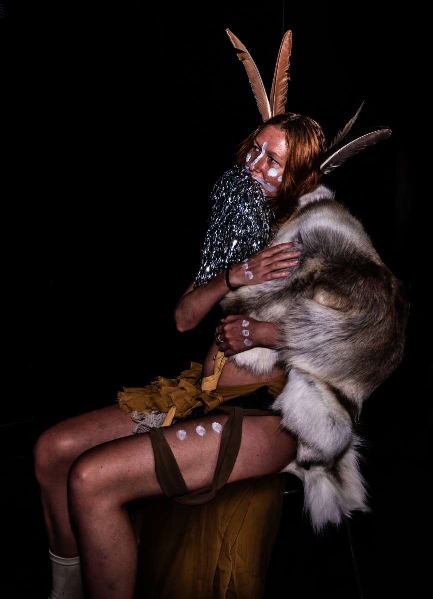 Seated in the dark embracing a reindeer skin, feathers rising from the hair
