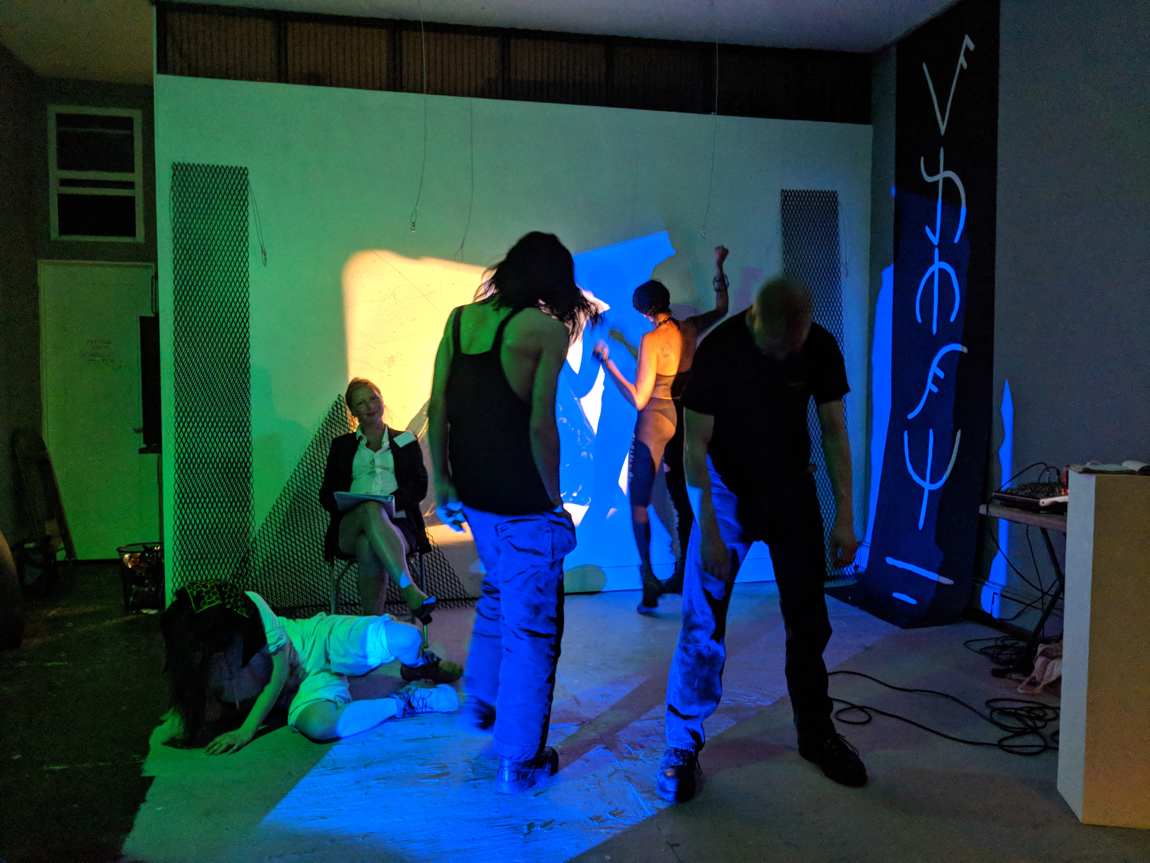 The ensemble moving across the space, a seated figure in a suit taking notes against the wall