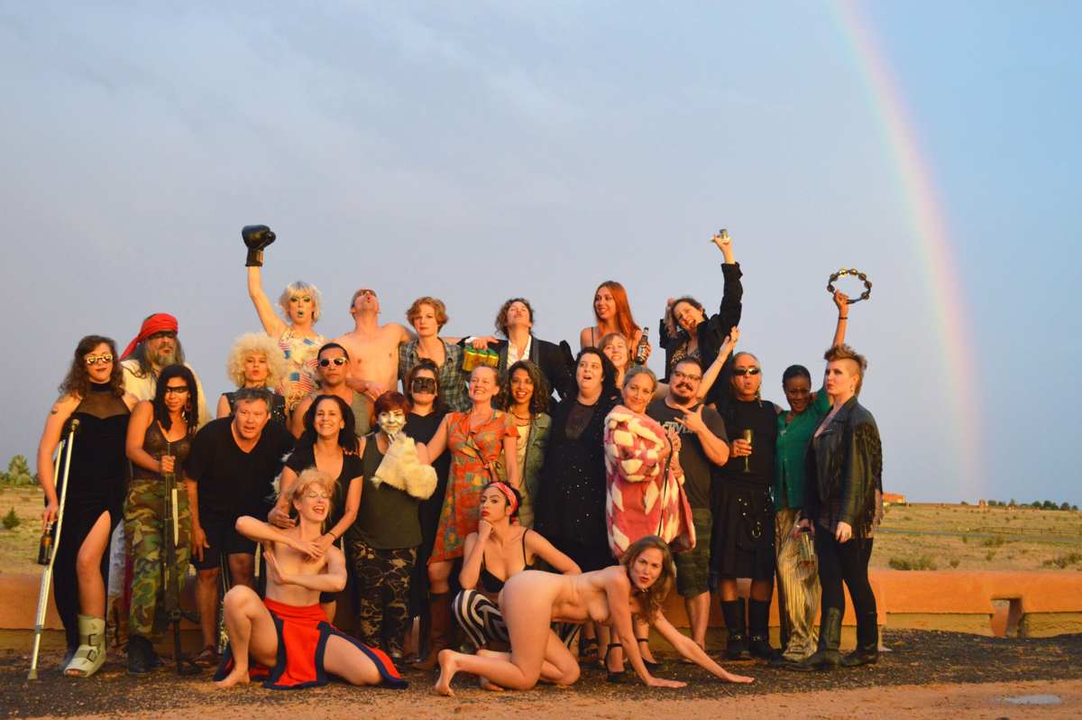 The full La Pocha Nostra workshop company posing in the New Mexico desert at golden hour, a rainbow arcing behind them
