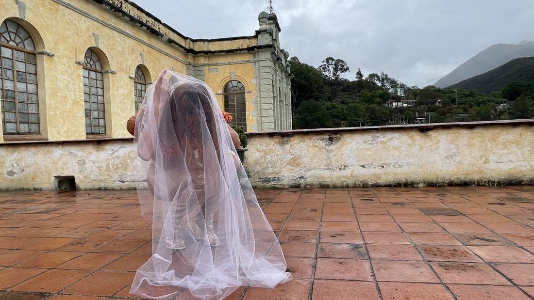 Two figures under one long tulle veil on a rain-wet rooftop terrace, yellow colonial building and green mountains behind — La Pocha Nostra workshop, Oaxaca