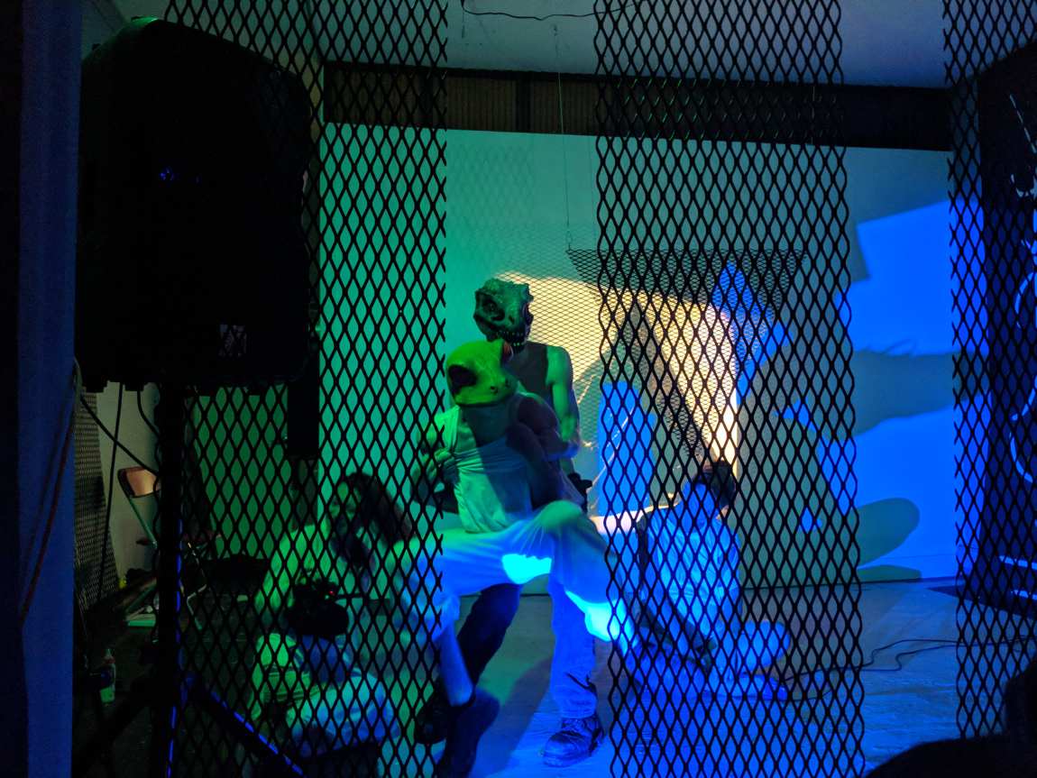 Two performers in reptilian masks embracing behind a black metal-lattice screen under green and blue light