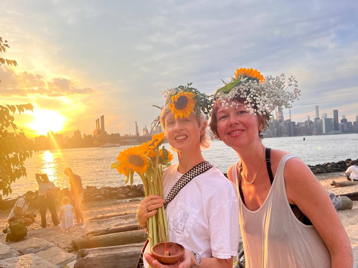 Two women in sunflower and baby's breath crowns holding sunflowers and a clay lamp on the beach at golden hour, Manhattan behind